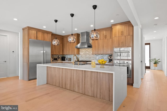 kitchen featuring pendant lighting, light hardwood / wood-style flooring, stainless steel appliances, a center island with sink, and wall chimney exhaust hood