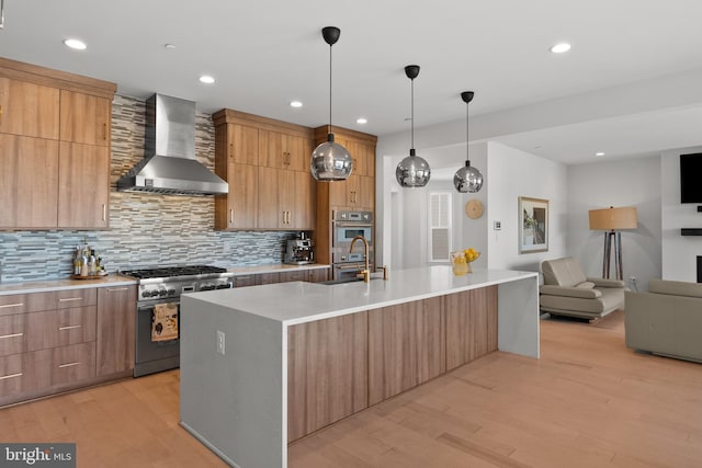 kitchen with appliances with stainless steel finishes, wall chimney exhaust hood, light hardwood / wood-style floors, decorative light fixtures, and a large island with sink