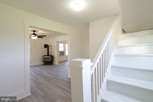 staircase with cooling unit, wood finished floors, a ceiling fan, baseboards, and a wood stove