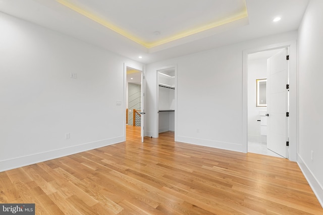 unfurnished bedroom featuring ensuite bathroom, a spacious closet, a raised ceiling, and light wood-type flooring