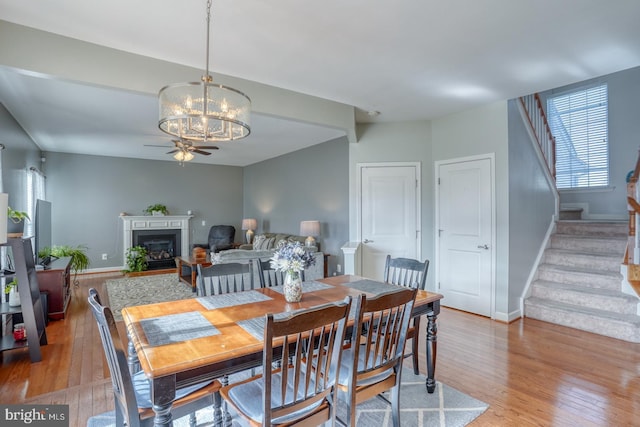 dining room featuring light wood-style flooring, a fireplace, stairway, and ceiling fan with notable chandelier