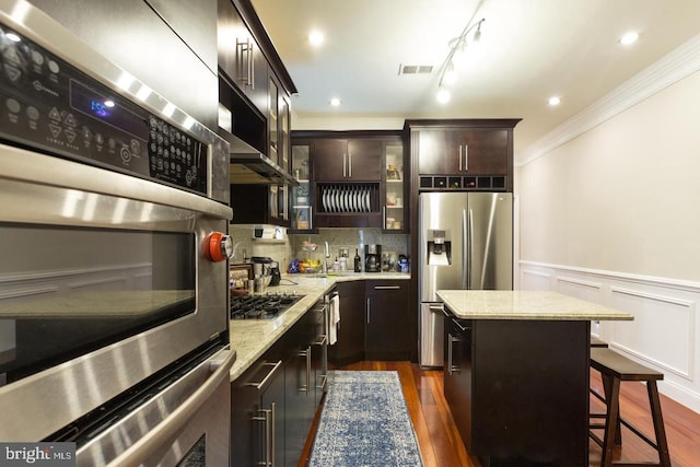 kitchen with crown molding, dark brown cabinets, stainless steel appliances, light stone counters, and a kitchen island