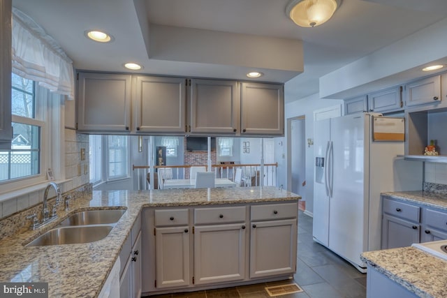 kitchen featuring sink, white refrigerator with ice dispenser, kitchen peninsula, light stone countertops, and decorative backsplash