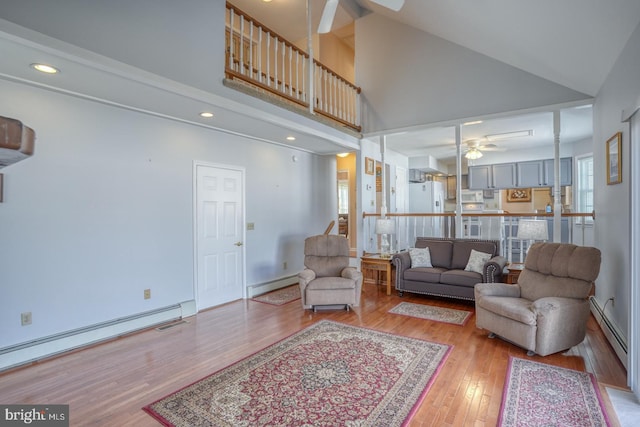 living room featuring baseboard heating, ceiling fan, light hardwood / wood-style floors, and a towering ceiling