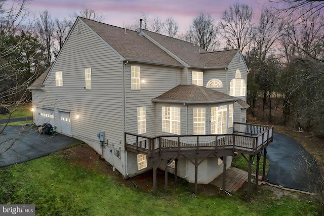 back of house at dusk with a garage, a deck, roof with shingles, and aphalt driveway