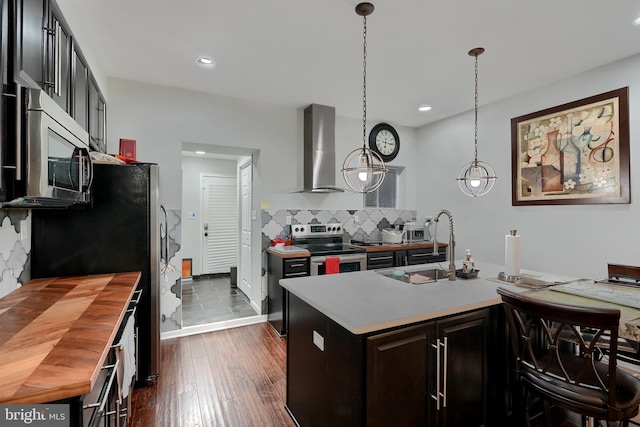 kitchen with pendant lighting, decorative backsplash, stainless steel appliances, a center island with sink, and wall chimney exhaust hood