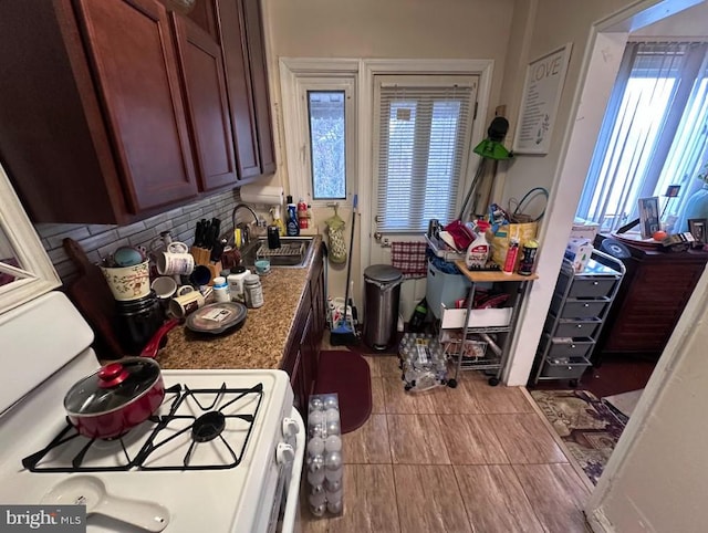 kitchen featuring sink, light tile patterned floors, white range with gas cooktop, dark stone counters, and backsplash