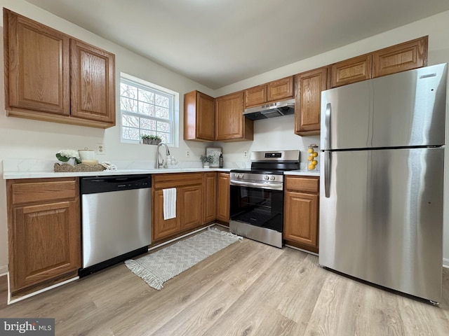 kitchen with brown cabinetry, light wood-style flooring, appliances with stainless steel finishes, light countertops, and under cabinet range hood