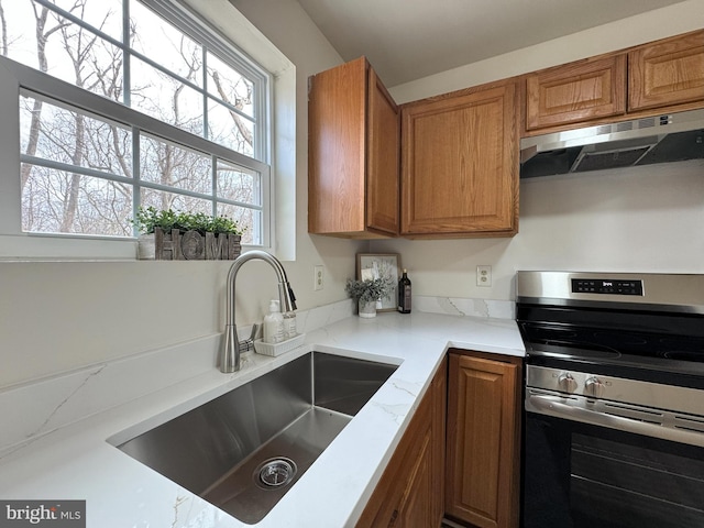 kitchen with light stone counters, brown cabinets, stainless steel electric range oven, a sink, and under cabinet range hood