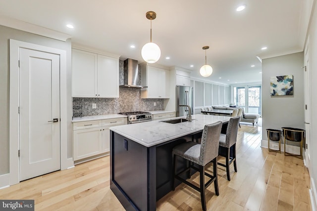 kitchen with an island with sink, sink, hanging light fixtures, light stone counters, and wall chimney exhaust hood