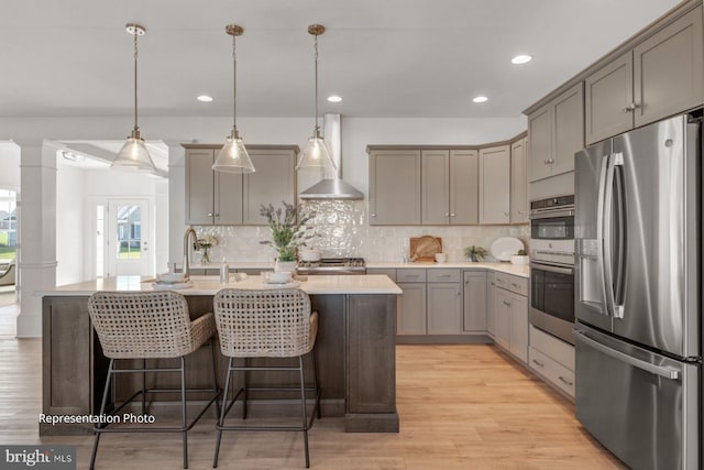 kitchen with sink, decorative light fixtures, a center island with sink, stainless steel appliances, and wall chimney range hood