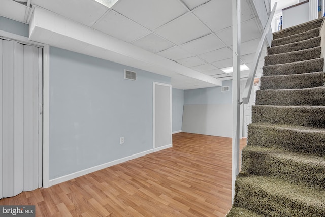 finished below grade area featuring visible vents, a paneled ceiling, light wood-style flooring, and stairs