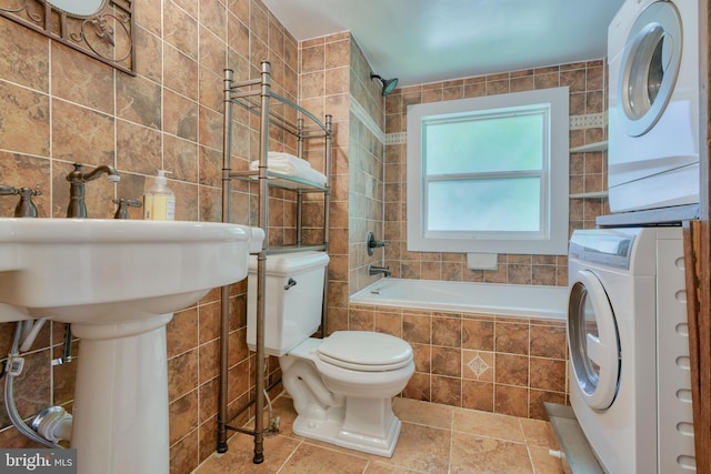 bathroom with stone tile floors, stacked washer / dryer, toilet, tiled shower / bath, and tile walls