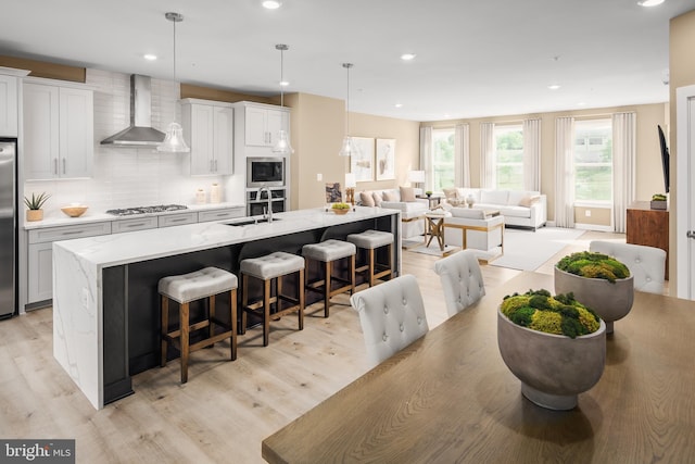 kitchen featuring wall chimney exhaust hood, pendant lighting, stainless steel appliances, a kitchen island with sink, and backsplash