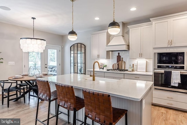 kitchen featuring custom exhaust hood, decorative light fixtures, appliances with stainless steel finishes, light stone countertops, and a kitchen island with sink