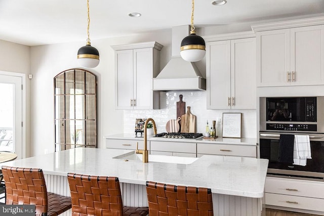 kitchen with a breakfast bar, custom exhaust hood, a kitchen island with sink, light stone counters, and stainless steel appliances