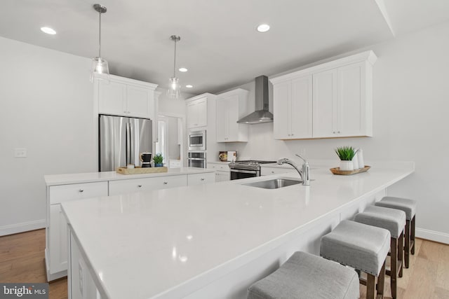 kitchen with wall chimney exhaust hood, appliances with stainless steel finishes, a peninsula, light wood-style floors, and a sink