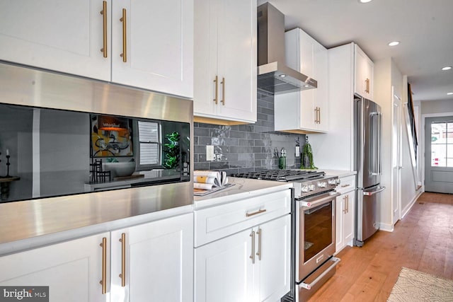 kitchen with wall chimney range hood, light hardwood / wood-style flooring, appliances with stainless steel finishes, backsplash, and white cabinets