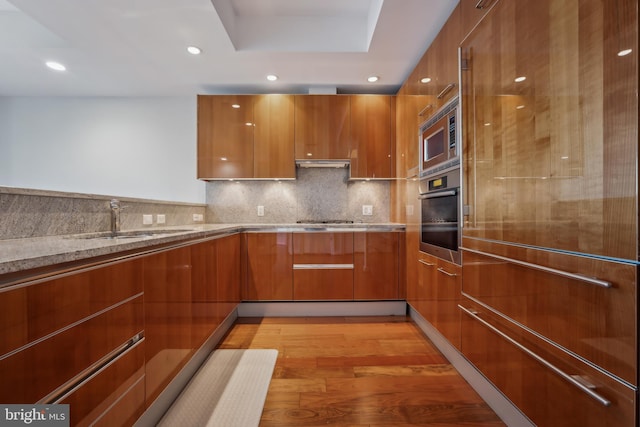 kitchen with sink, light hardwood / wood-style flooring, range hood, built in appliances, and tasteful backsplash