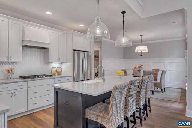 kitchen with decorative light fixtures, appliances with stainless steel finishes, wall chimney range hood, a kitchen island with sink, and white cabinets