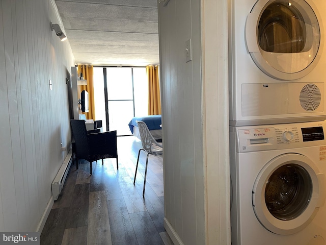 laundry room featuring baseboard heating, wooden walls, stacked washing maching and dryer, and dark hardwood / wood-style flooring