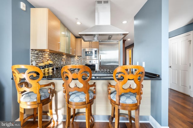 kitchen featuring light brown cabinetry, built in appliances, dark hardwood / wood-style floors, island exhaust hood, and backsplash