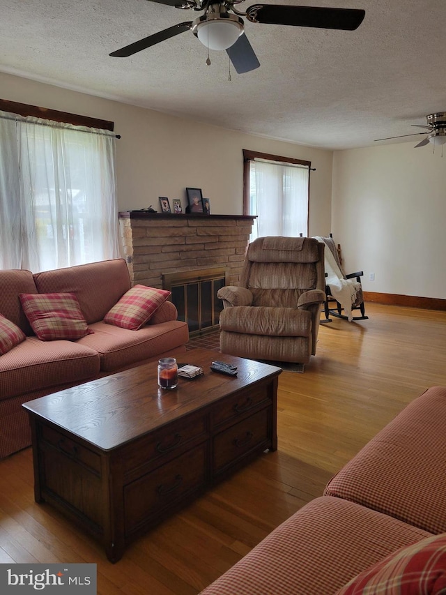 living room with ceiling fan, a stone fireplace, light hardwood / wood-style flooring, and a textured ceiling