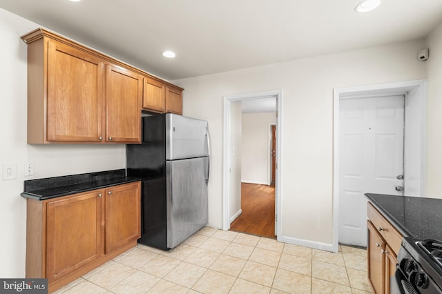 kitchen with light tile patterned floors, freestanding refrigerator, brown cabinets, dark stone countertops, and range with gas cooktop