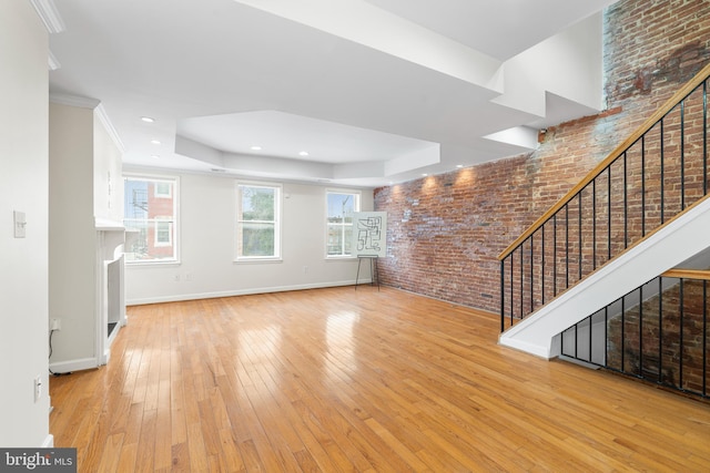 unfurnished living room with baseboards, brick wall, stairway, hardwood / wood-style floors, and a tray ceiling