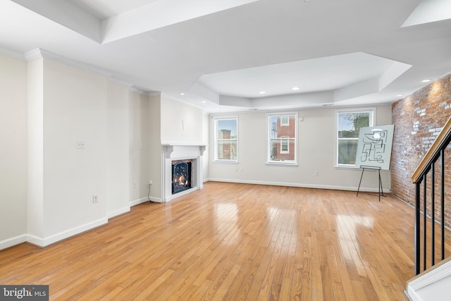 unfurnished living room with a tray ceiling, light wood-style floors, a warm lit fireplace, baseboards, and stairs