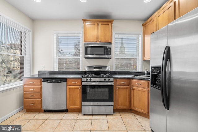 kitchen featuring dark countertops, appliances with stainless steel finishes, brown cabinetry, light tile patterned flooring, and a sink