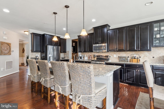 kitchen featuring hanging light fixtures, appliances with stainless steel finishes, light stone countertops, a kitchen island with sink, and decorative backsplash