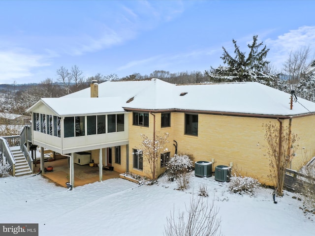 snow covered property featuring central air condition unit, a sunroom, stairs, a carport, and a chimney