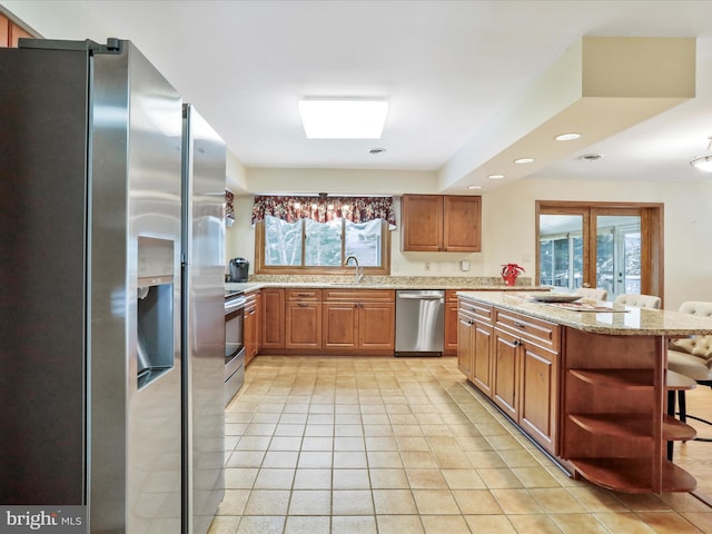 kitchen with brown cabinetry, light stone counters, stainless steel appliances, open shelves, and a sink