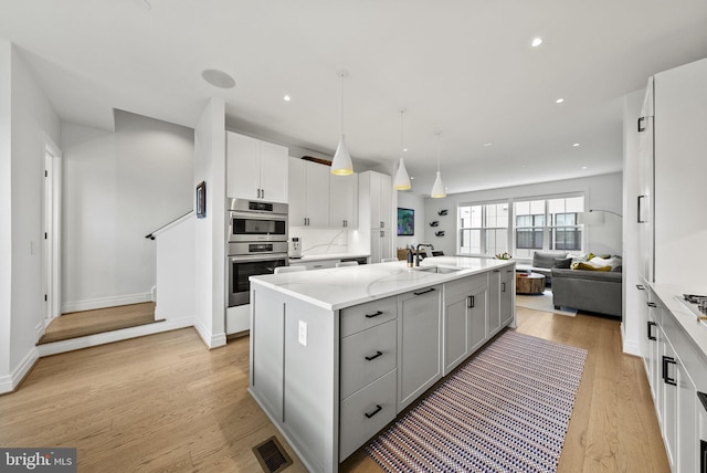 kitchen with double oven, an island with sink, sink, hanging light fixtures, and light stone countertops