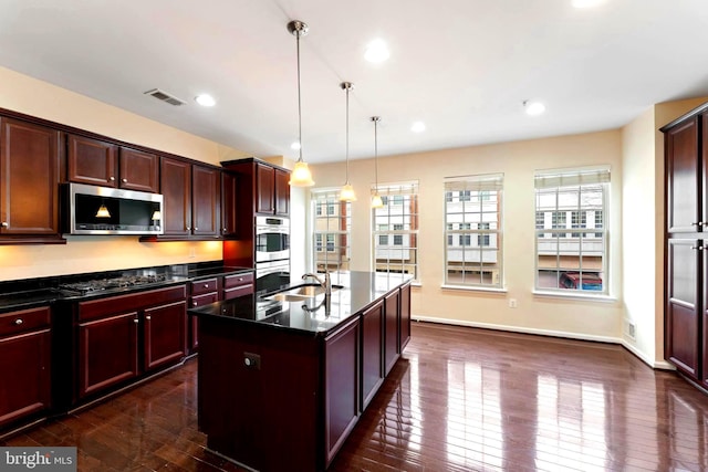 kitchen featuring dark hardwood / wood-style floors, decorative light fixtures, sink, stainless steel appliances, and a center island with sink