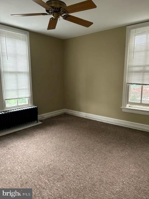 carpeted empty room featuring radiator, a ceiling fan, baseboards, and a wealth of natural light