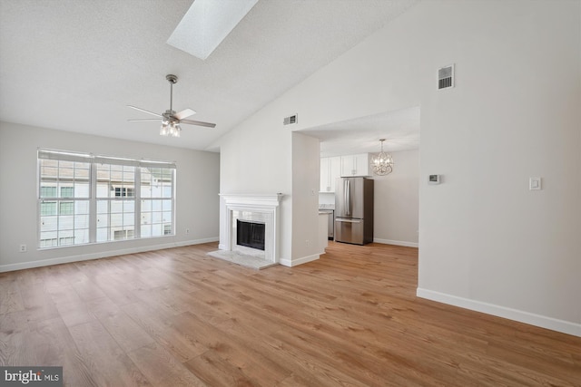 unfurnished living room featuring ceiling fan with notable chandelier, high vaulted ceiling, a skylight, a textured ceiling, and light hardwood / wood-style flooring