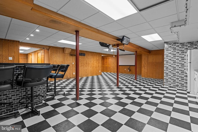 basement with white refrigerator, bar, a drop ceiling, and wood walls