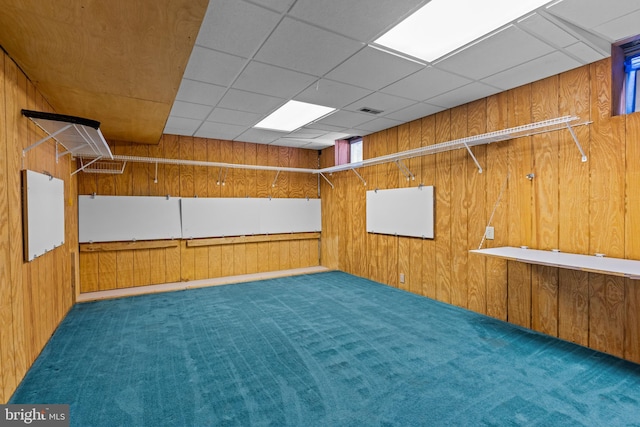 basement featuring dark colored carpet, a paneled ceiling, and wooden walls