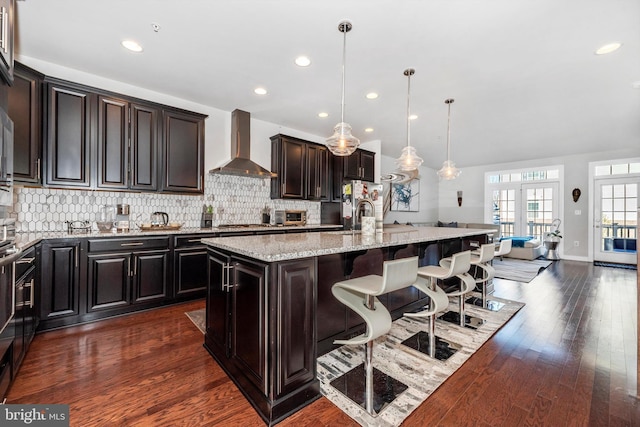 kitchen with a kitchen bar, decorative light fixtures, an island with sink, light stone countertops, and wall chimney range hood