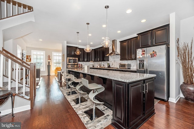 kitchen with a large island with sink, appliances with stainless steel finishes, pendant lighting, wall chimney range hood, and backsplash