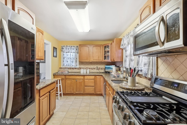 kitchen featuring light stone counters, a sink, appliances with stainless steel finishes, backsplash, and glass insert cabinets