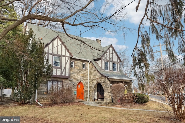 tudor-style house featuring fence, stone siding, roof with shingles, stucco siding, and a chimney