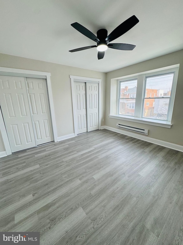 unfurnished bedroom with two closets, light wood-type flooring, ceiling fan, and baseboard heating