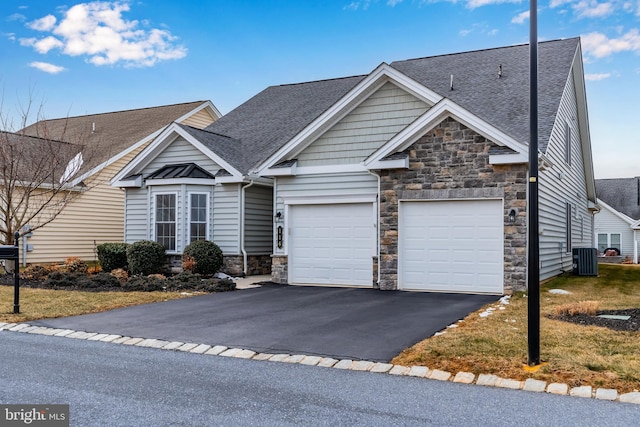 view of front of home featuring an attached garage, roof with shingles, central AC unit, stone siding, and driveway