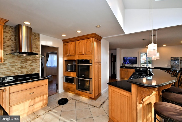 kitchen with decorative light fixtures, double oven, wall chimney range hood, a kitchen island with sink, and black stovetop