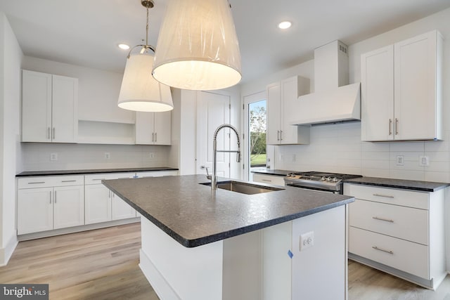 kitchen featuring white cabinetry, sink, hanging light fixtures, wall chimney range hood, and a center island with sink