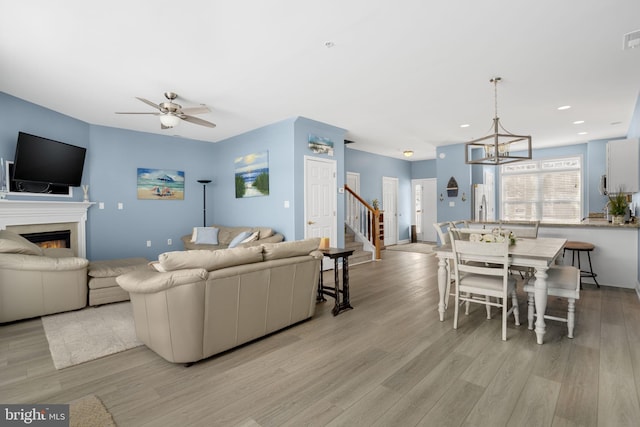 living room with a glass covered fireplace, light wood-style flooring, stairs, ceiling fan with notable chandelier, and recessed lighting