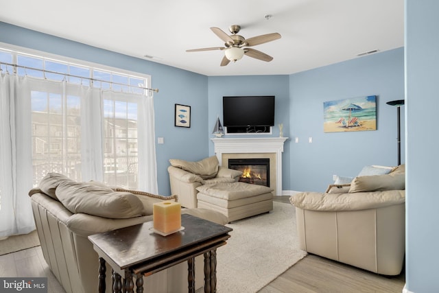 living area with light wood-type flooring, a glass covered fireplace, visible vents, and a ceiling fan